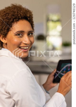 Woman turning toward camera in kitchen lab space, wearing white lab coat, holding tablet with graph Woman turning toward camera in kitchen lab space, wearing white lab coat, holding tablet with graph 128740129