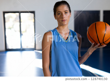Female basketball player standing on gym court, holding basketball by glass doors, copy space 128740130