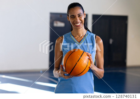 Woman standing on indoor court wearing light blue jersey, holding orange basketball and smiling Woman standing on indoor court wearing light blue jersey, holding orange basketball and smiling 128740131