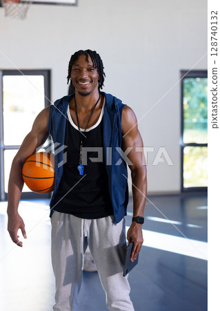 African American man standing in basketball gym, smiling and holding basketball, tablet and whistle African American man standing in basketball gym, smiling and holding basketball, tablet and whistle 128740132
