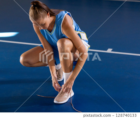 Female athlete tying sneaker laces on indoor court, wearing pastel sneakers and blue jersey Female athlete tying sneaker laces on indoor court, wearing pastel sneakers and blue jersey 128740133
