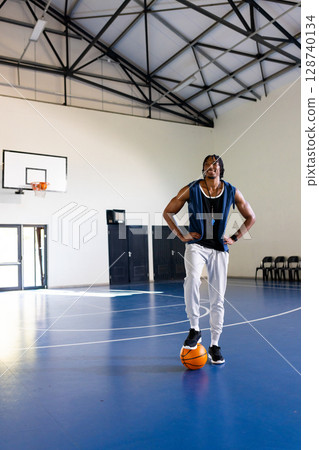 African American man standing with foot on basketball in indoor court, wearing whistle, copy space 128740134