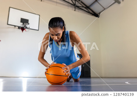 Diverse female athlete kneeling on gym floor pressing orange basketball under hoop, stretching core 128740139