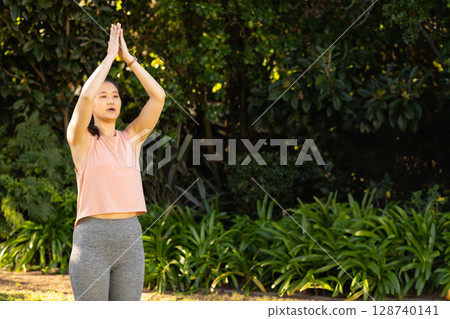Asian woman raising arms above head in lush garden, with fitness tracker and sunshine, copy space 128740141