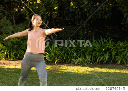 Mid-adult Asian woman practicing Warrior II yoga pose on grassy lawn, enjoying warm sunlight 128740145