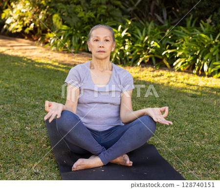 Senior Asian Chinese woman meditating on black yoga mat in grassy garden, practicing mudra pose 128740151