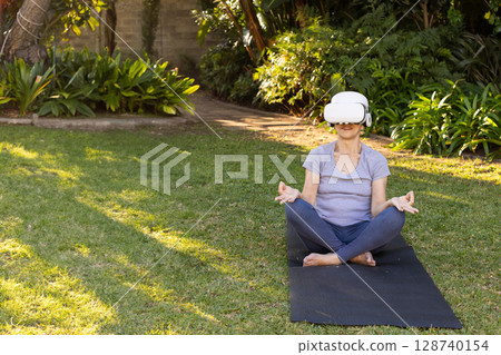 Senior Asian woman meditating on yoga mat in backyard, wearing VR headset, copy space Senior Asian woman meditating on yoga mat in backyard, wearing VR headset, copy space 128740154