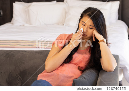 Asian woman sitting on grey sofa in bedroom, wiping face with tissue sadly, copy space 128740155