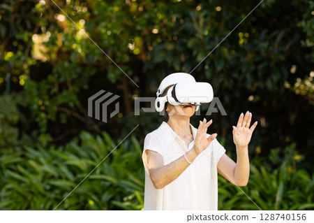 Senior Asian woman experiencing virtual reality in lush outdoor garden, with white VR headset 128740156