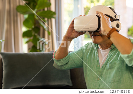 Middle-aged Asian man adjusting white VR headset on dark green sofa in living room, copy space 128740158