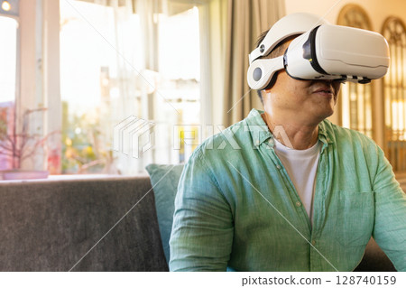 Asian senior man using VR headset on gray sofa near windows in living room, copy space Asian senior man using VR headset on gray sofa near windows in living room, copy space 128740159