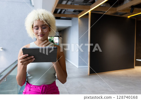 Woman standing in modern office corridor leaning against glass railing, holding tablet and reading 128740168
