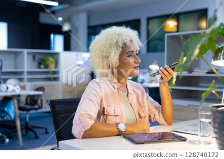 African American woman speaking into smartphone at desk in office, with laptop and tablet African American woman speaking into smartphone at desk in office, with laptop and tablet 128740172