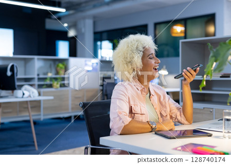 Mid-adult man speaking into smartphone in open-plan office at desk, with laptop, tablet, copy space Mid-adult man speaking into smartphone in open-plan office at desk, with laptop, tablet, copy space 128740174