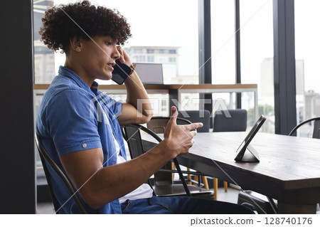 Curly-haired man talking on smartphone at conference table in office space, with tablet and laptop Curly-haired man talking on smartphone at conference table in office space, with tablet and laptop 128740176