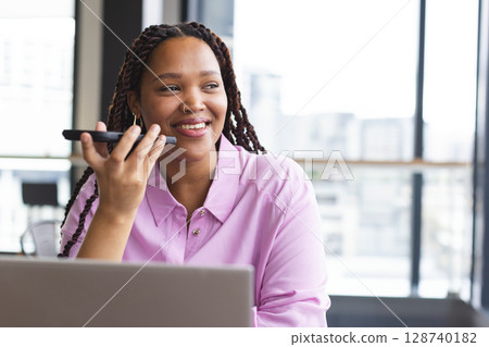 Diverse woman speaking into smartphone, working on laptop in office, with city skyline, copy space Diverse woman speaking into smartphone, working on laptop in office, with city skyline, copy space 128740182