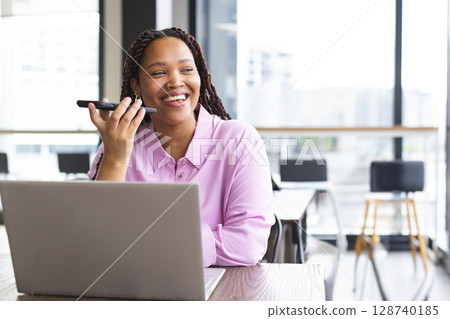 Woman speaking into smartphone while working on laptop at open plan office with desk, copy space 128740185