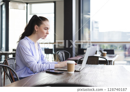 Woman typing on silver laptop at modern cafe with large windows, holding coffee cup and smartphone 128740187