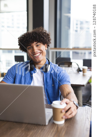 Young man working on laptop at modern cafe table, with headphones and holding paper coffee cup 128740188