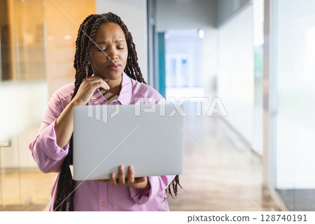 Multiracial young woman standing in modern office hallway, holding laptop and pondering ideas Multiracial young woman standing in modern office hallway, holding laptop and pondering ideas 128740191
