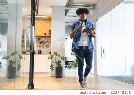 Multiracial man walking through office kitchen area, with tablet, headphones and plants, copy space Multiracial man walking through office kitchen area, with tablet, headphones and plants, copy space 128740196