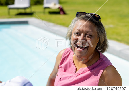 Relaxed senior woman enjoying sun on pool edge in backyard with sunglasses, copy space 128740207