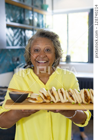 Senior woman presenting wooden serving board with steamed buns and dipping sauce in home kitchen Senior woman presenting wooden serving board with steamed buns and dipping sauce in home kitchen 128740214