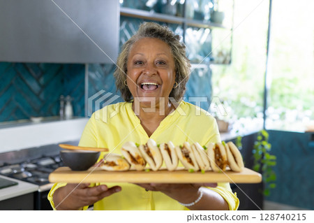Senior woman holding serving board with stuffed buns in modern kitchen, offering sauce bowl 128740215