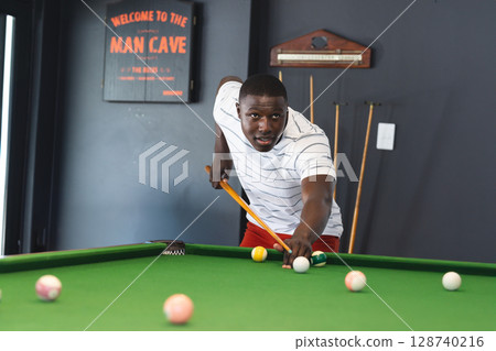 Young African American man shooting pool on green pool table in man cave, with cue stick 128740216