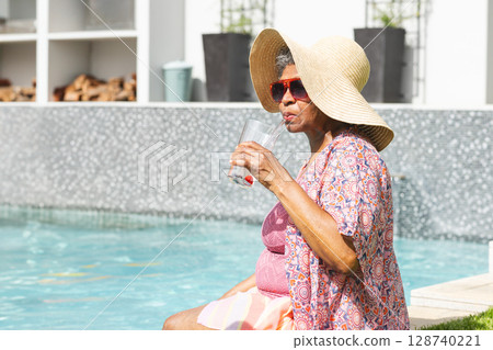 Senior woman sitting at poolside, wearing straw hat, red sunglasses, sipping from glass, copy space 128740221