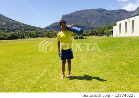 Boy standing barefoot on bright green lawn beside white building, holding blue yoga mat 128740225