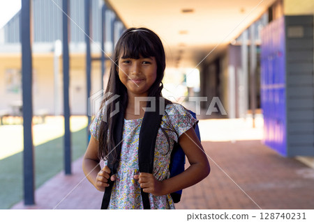 8-year-old girl wearing floral dress standing in sunlit school corridor, gripping blue backpack 128740231
