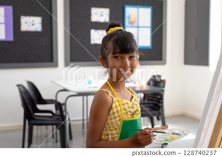 7-year-old girl painting on canvas at easel with palette and brush in art classroom, copy space 128740237