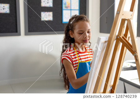 Female child painting on canvas on easel in art classroom, with paintbrush and pinned drawings 128740239