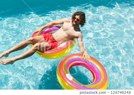 Young man floating on multicolored inflatable ring in outdoor swimming pool, holding second ring 128740248