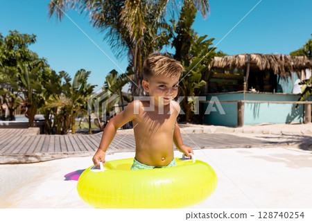 Boy child standing in tropical resort pool, wearing green swim trunks and holding yellow swim ring 128740254