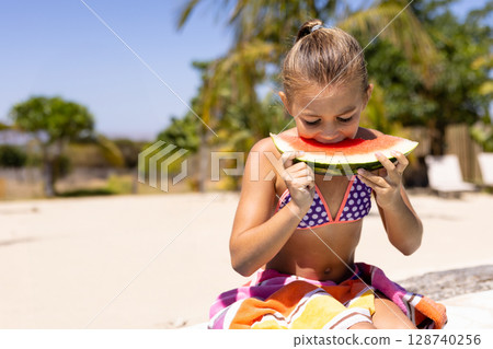 Child girl biting large watermelon slice while sitting on colorful towel on bench in backyard Child girl biting large watermelon slice while sitting on colorful towel on bench in backyard 128740256