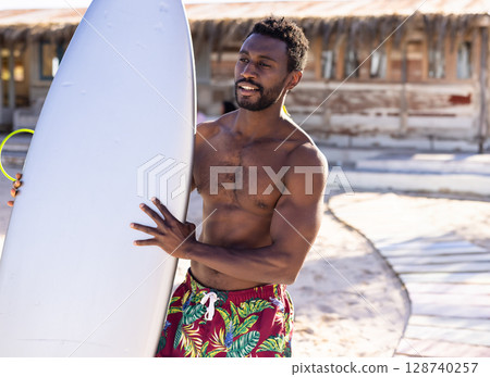 African American man on sandy beach near shack holding white surfboard, wearing red trunks 128740257