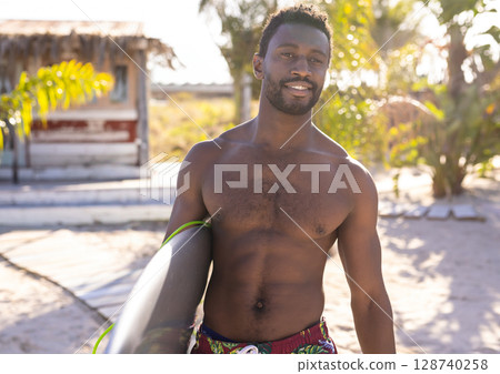Young adult African American man standing on tropical beach holding surfboard, enjoying sunshine 128740258