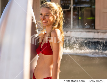 Young adult woman wearing red bikini standing in shallow water by pier, holding white surfboard 128740259