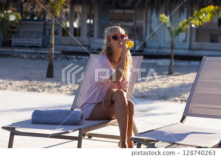 Woman sitting on lounge chair at beach patio, wearing sunglasses and sipping orange juice 128740263