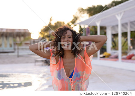 African American woman raising arms on boardwalk at sunset, wearing orange cover-up and bikini top 128740264