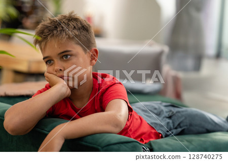 Young boy reclining on green sofa with pillows in living room, displaying pensive expression Young boy reclining on green sofa with pillows in living room, displaying pensive expression 128740275
