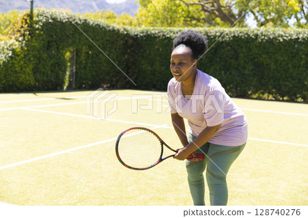 Mature African American woman taking ready stance on tennis court holding tennis racket, copy space 128740276