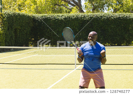 Senior African American man pumping fist, holding tennis racket on outdoor tennis court, copy space 128740277