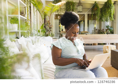 Mature African American woman typing on laptop in sunroom, with couch, hanging plants, copy space 128740286