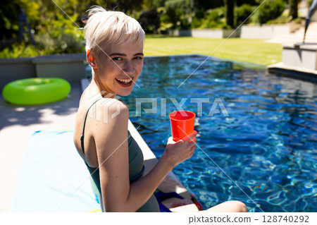 non-binary person sitting on home pool deck holding red cup beside blue towel and neon-green ring 128740292