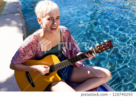 Non-binary person playing acoustic guitar at pool edge, sparkling water reflections, copy space 128740293