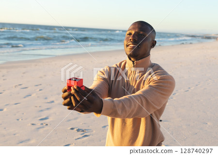 African American man kneeling on sandy shore, presenting red box holding diamond ring, copy space 128740297