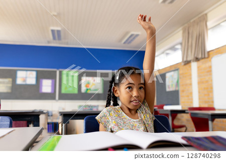 Young African American girl raising hand at desk in elementary school classroom with open workbook 128740298